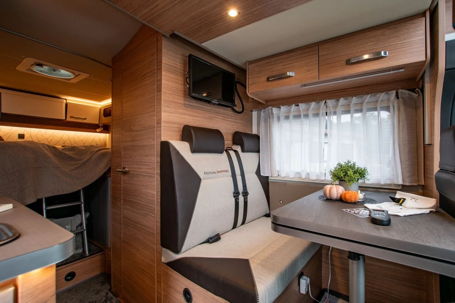モダンな木目調で統一されたキャンピングカーの車内です。二段ベッド、ダイニングスペース、テレビ、簡易キッチンが備え付けられており、快適な旅行を予感させる居住空間が広がっています。