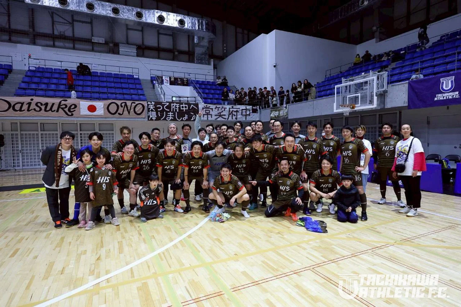 フットサルチーム「TACHIKAWA ATHLETIC F.C.」の選手と子供、関係者が体育館のコートで集合写真を撮っている