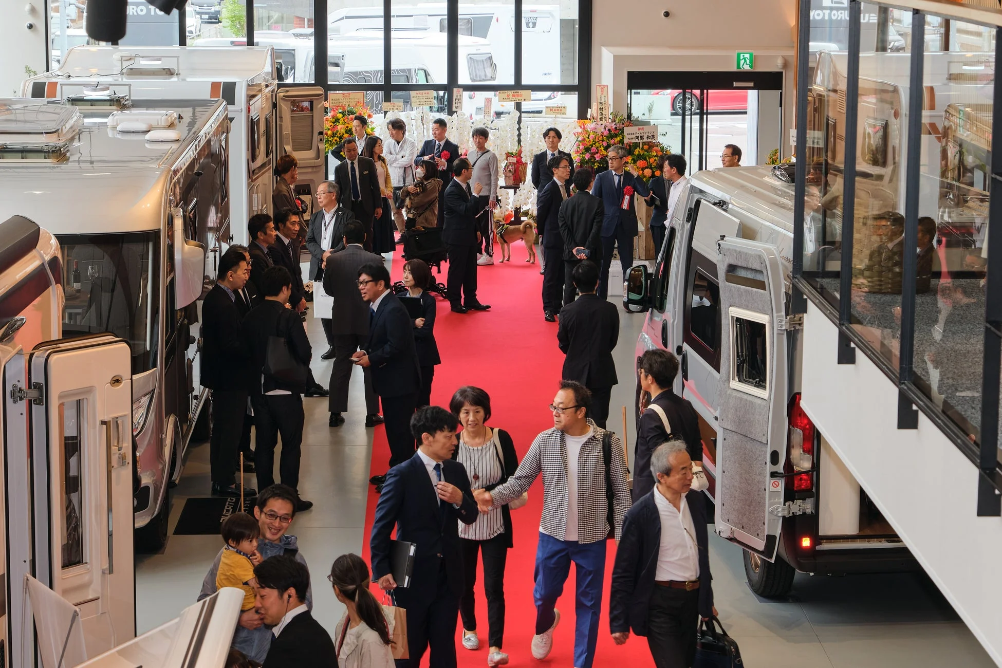 キャンピングカーのショールームで多くの人々が賑わうイベントの様子。赤絨毯が敷かれ、新型車両の展示やビジネス関係者、一般客、家族連れなどが交流している。