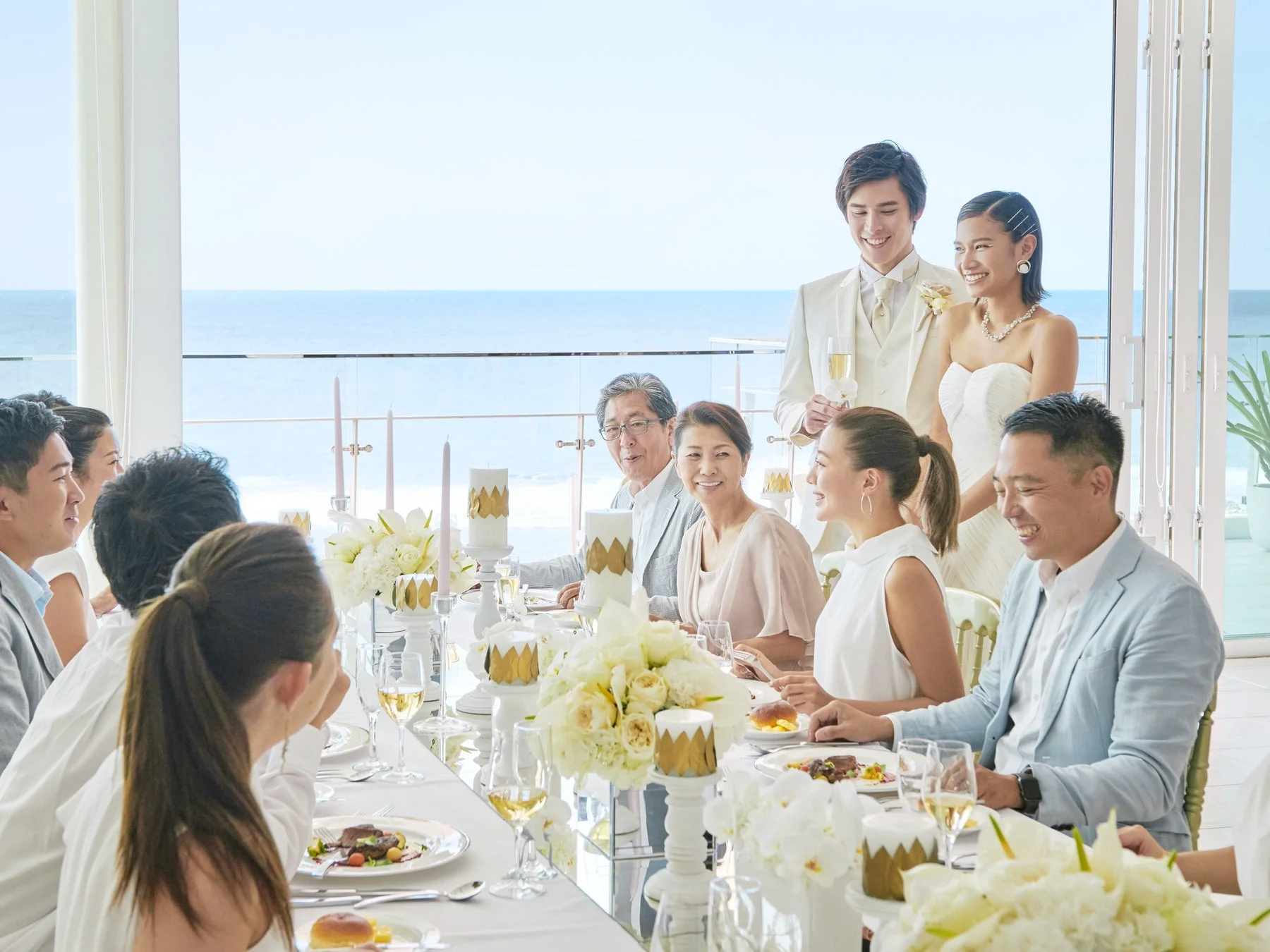海が見える明るい会場で、新郎新婦とゲストが笑顔で食事を楽しむ結婚披露宴の様子。