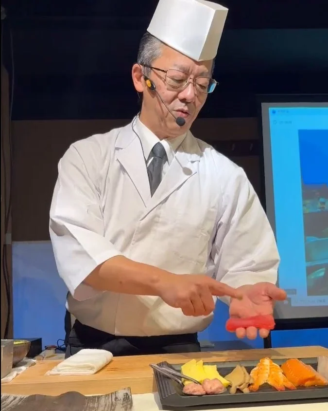 白い調理服の寿司職人がヘッドセットをつけ、手でマグロのネタを持ち、寿司の握り方を実演している