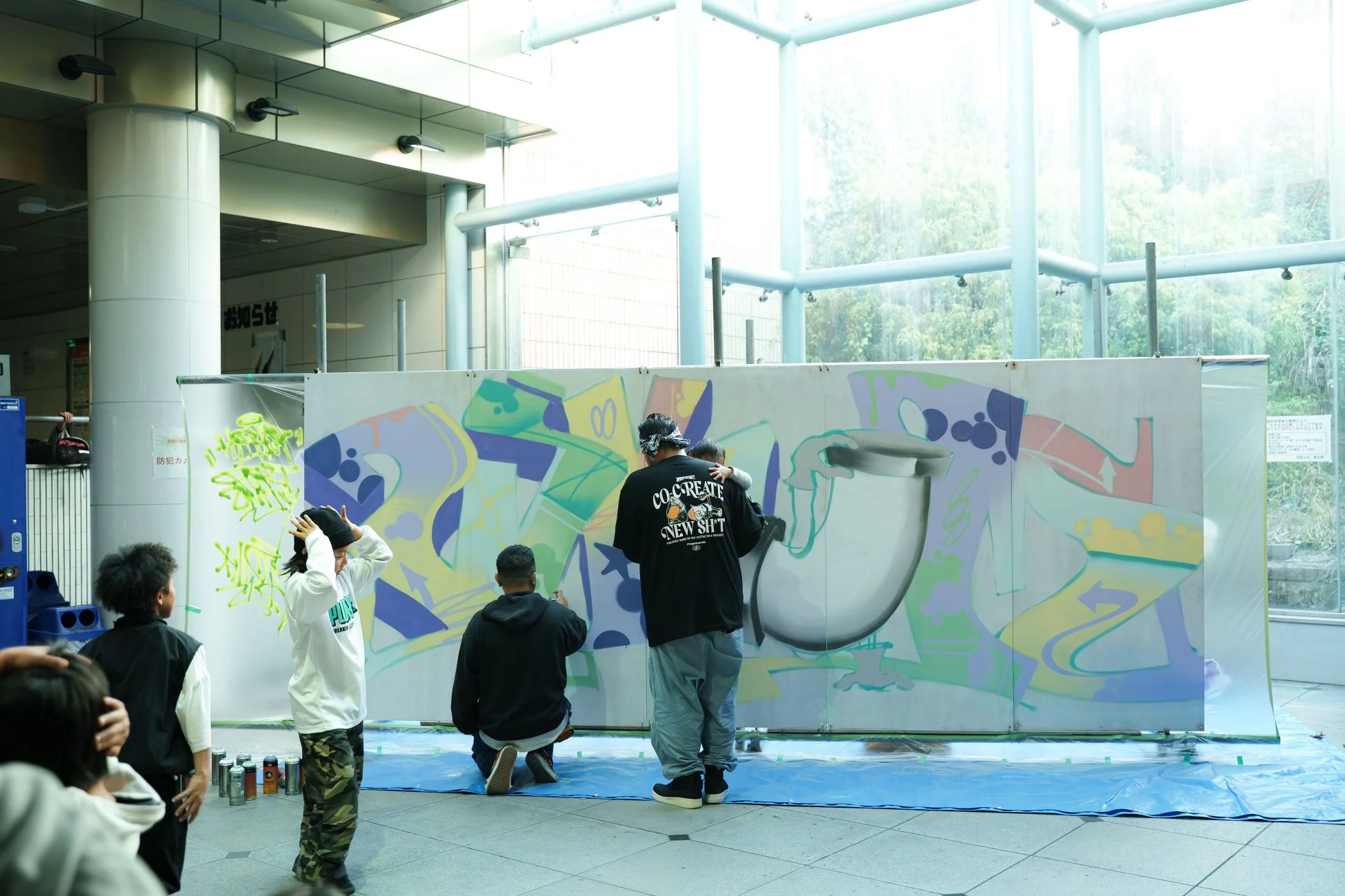 屋内空間で、大人と子供たちが協力して大きなキャンバスにスプレーアートを描いている様子