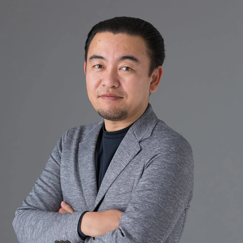 グレーの背景を背に、グレーのジャケットとダークカラーのインナーを着用し、腕を組んでカメラを見つめるアジア人男性のバストアップポートレート。落ち着いた表情で信頼感を醸し出しています。