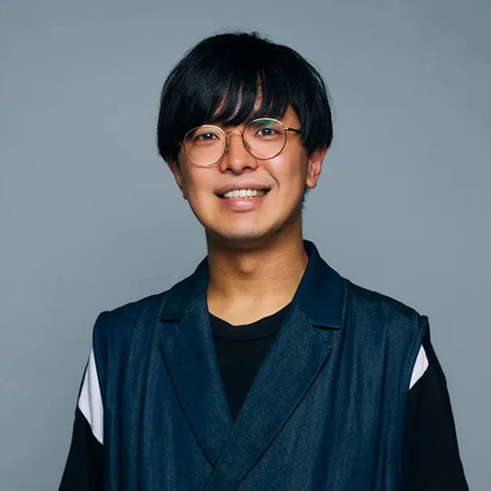 笑顔でカメラを見つめる丸眼鏡をかけた若い日本人男性のポートレートです。黒いTシャツの上に紺色のベストを着用しており、背景はシンプルなグレーです。